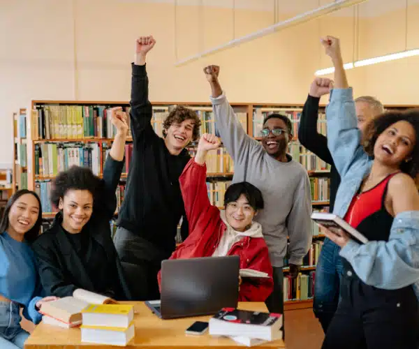 Groupe de collégiens et lycéens enthousiastes levant les bras en l'air dans une bibliothèque, lors d'une activité pédagogique collaborative
